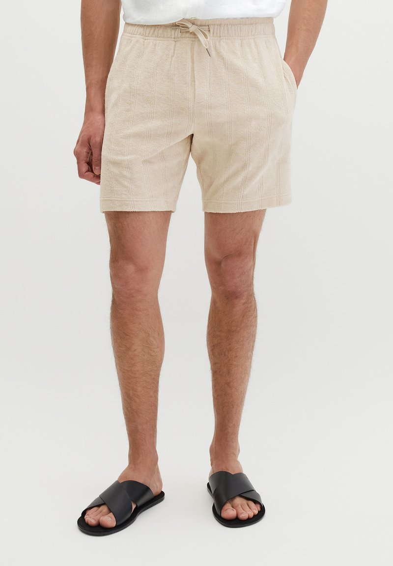 Uomo che indossa pantaloncini beige strutturati con coulisse e tasche laterali e sandali neri con cinturini incrociati su sfondo semplice.