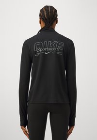Crna dugorukava atletska majica s logotipom "NIKE Sportswear" otisnutim sivo na leđima, s teksturiranim materijalom i uskim krojem.