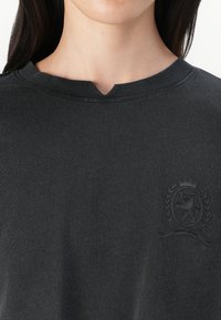 Mørk grå, T-skjorte med rund hals laget av myk stoff, med en subtil preget logo på venstre bryst. Korte ermer og en enkel design.