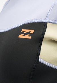 Κοντινή λήψη μαύρης, μπεζ και ανοιχτό μωβ στολής καταδύσεων με πορτοκαλί λογότυπο κύματος και ορατές ραφές.