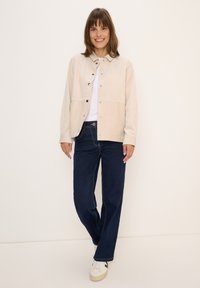 Vrouw die glimlacht, gekleed in een beige knoop-jes corduroy jas, witte shirt, donkerblauwe spijkerbroek en witte sneakers, staande tegen een lichte achtergrond.