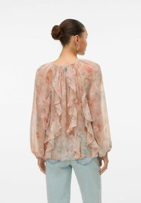 Blusa in chiffon floreale con maniche lunghe, dettaglio arricciato sul retro, colore pesca chiaro e vestibilità ampia, abbinata a jeans azzurri chiari.