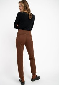 Brązowe spodnie z wysokim stanem o gładkiej fakturze i subtelnym wykończeniu kieszeni z tyłu, zestawione z czarną bluzką z długim rękawem i masywnymi czarnymi butami.