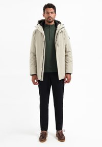 Beige isolerende jas met een capuchon over een donkergroene trui, gecombineerd met zwarte broek en bruine schoenen; heeft een ritszak en een gladde textuur.