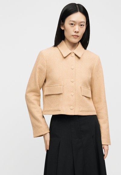 Femme portant une veste beige à motif pied-de-poule boutonnée avec poches à rabats, associée à une jupe noire taille haute sur un fond uni.