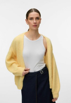 Kvinne iført en hvit tanktopp, en tykk gul cardigan og et mørkeblått skjørt med høy midje og et belte med sølvspenne.