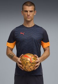 Koszulka sportowa w ciemnym granacie z pomarańczowymi rękawami i wzorem w zygzak, trzymana ze złotą piłką nożną z wzorem i czerwoną logo.