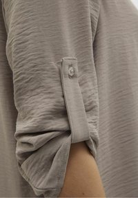 Camicia in tessuto grigio e testurizzato con maniche arrotolate e dettaglio a bottone, che mostra leggere pieghe e un aspetto morbido.