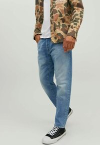 Camisa de camuflaje en tonos marrones y verdes, camiseta blanca debajo, combinada con vaqueros de algodón azul claro y zapatillas negras de corte bajo.