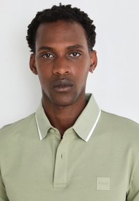 Polo shirt vert clair avec un col structuré, des bords blancs et un patch de logo carré sur la poitrine gauche. Texture de tissu douce.