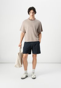 Tricou din bumbac stripat maro și bej, pantaloni scurți bleumarin, adidași albi și gri, purtând o geantă tote bej. Design simplu și relaxat.