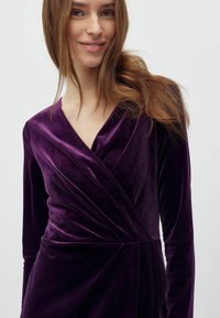 Violetinis aksominis suknelė ilgomis rankovėmis, V formos iškirpte ir drapiruotais detalėmis. Minkšta tekstūra ir elegantiška siluetas, tinkama proginėms iškilmėms.