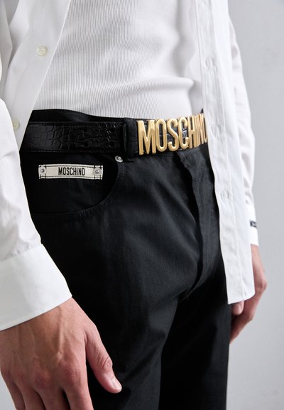 Must mustriba kuldsete "MOSCHINO" tähtedega ja krokodilli tekstuuriga. Ühendatud mustade pükstega ja valge särgiga, millel on Moschino silt.