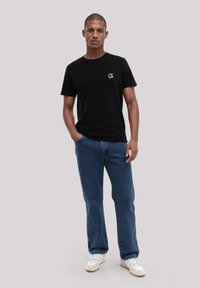 Schwarzes Baumwoll-T-Shirt mit einem kleinen grafischen Detail, kombiniert mit blauen Denim-Jeans und weißen Sneakers. Das Modell steht in entspannter Haltung.