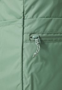 Tessuto in nylon verde menta con texture trapuntata. Presenta una piccola tasca con zip e un tirante a toggle, oltre a un dettaglio di cucitura rinforzata.