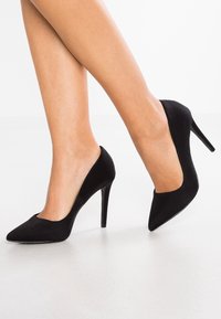 Chaussures à talons hauts noires avec une texture en daim, un bout pointu et des côtés découpés. Le talon est fin et mesure plusieurs centimètres de haut.