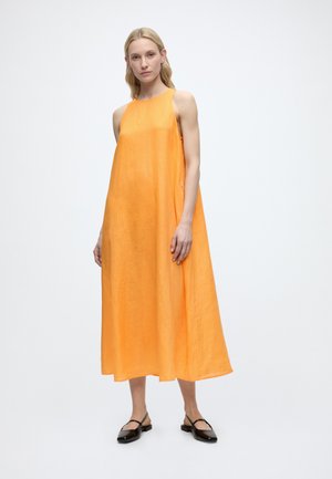 Vrouw staat in een mouwloze, losvallende oranje midi-jurk en zwarte platte schoenen tegen een effen witte achtergrond.