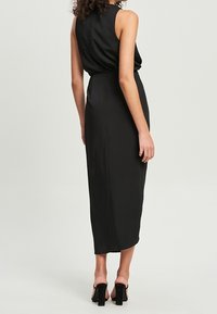 Vestido midi negro sin mangas con silueta ajustada, tela suave y detalle de cintura fruncida. Lleva puestas unas gruesas tacones negros.
