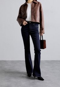 Veste en cuir marron courte portée sur une chemise blanche, associée à un jean évasé bleu foncé taille haute et un petit sac à main marron.