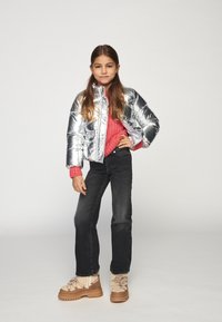 Νεαρή κοπέλα ποζάρει με το χέρι στη μέση, φορώντας ασημένιο μπουφάν, φωτεινό ροζ πουλόβερ, μαύρα τζιν και ταμπά χειμερινά μποτάκια, μπροστά σε απλό φόντο.