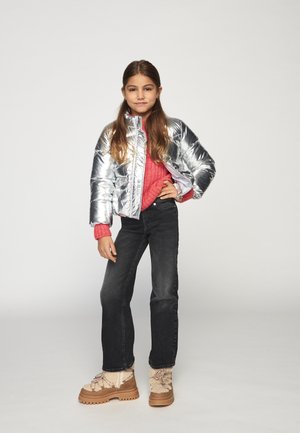 Fiatal lány pózolva, kezét a csípőjén pihentetve, ezüst színű pufi dzsekiben, élénk rózsaszín pulóverben, fekete farmernadrágban és bézs téli csizmában áll egy sima háttér előtt.