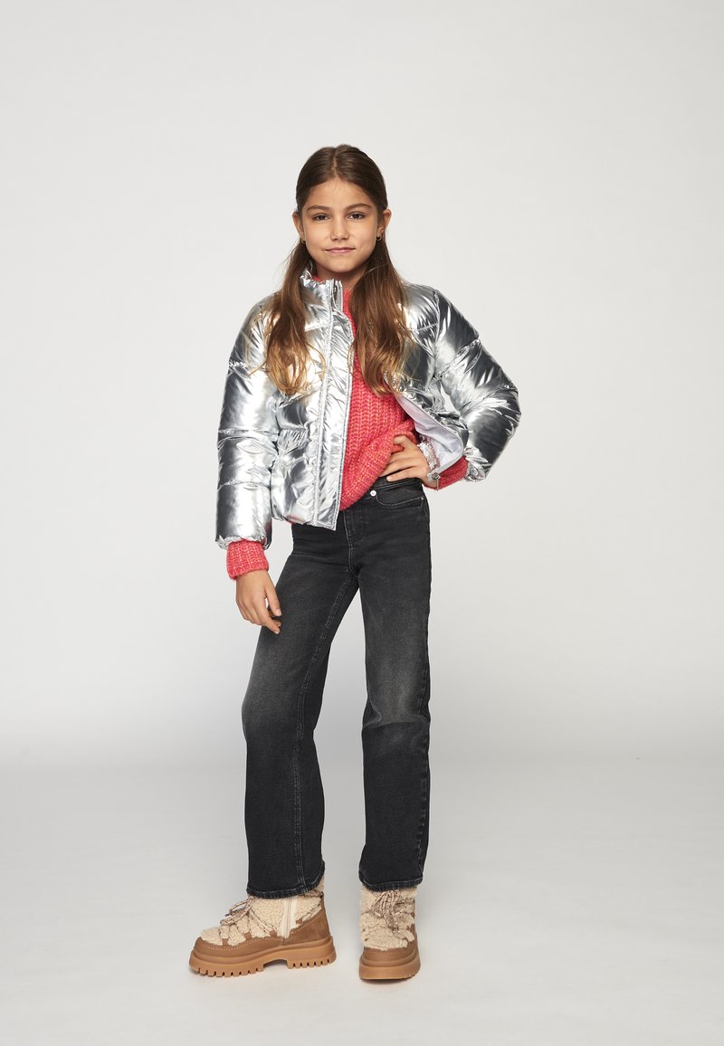 Νεαρή κοπέλα ποζάρει με το χέρι στη μέση, φορώντας ασημένιο μπουφάν, φωτεινό ροζ πουλόβερ, μαύρα τζιν και ταμπά χειμερινά μποτάκια, μπροστά σε απλό φόντο.