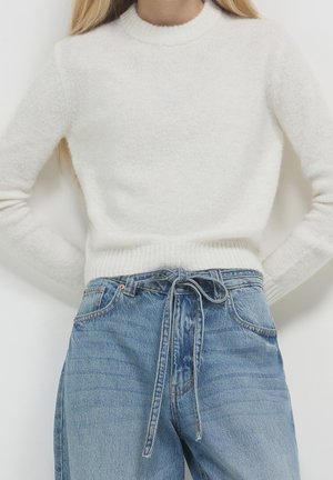 Femme portant un pull blanc duveteux rentré dans un jean bleu taille haute, ceinturé à la taille avec une ceinture en denim assortie.