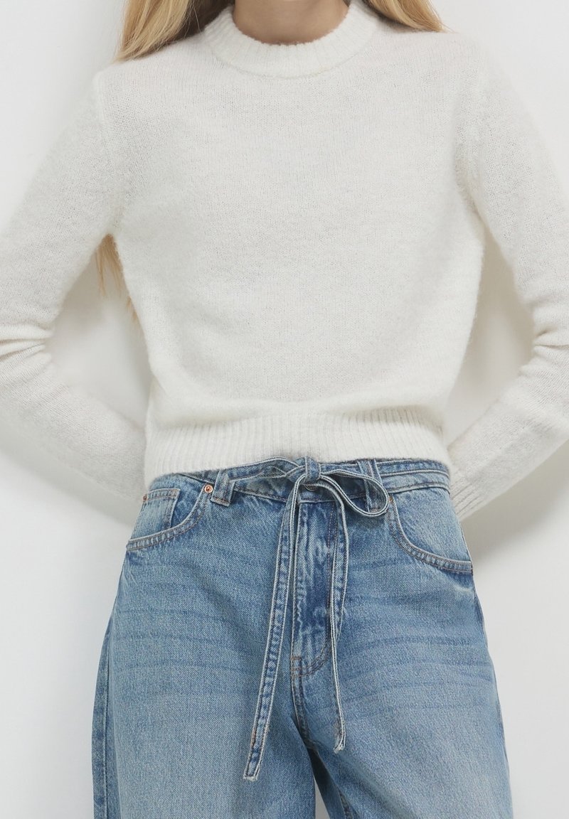 Femme portant un pull blanc duveteux rentré dans un jean bleu taille haute, ceinturé à la taille avec une ceinture en denim assortie.