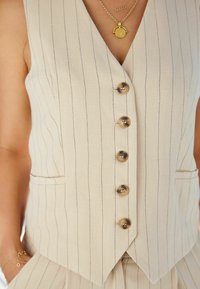 Gilet di lino beige con strisce verticali, dotato di cinque bottoni rotondi, vestibilità slim e due tasche laterali. Accentuato da gioielli dorati.