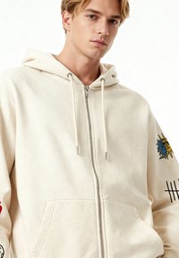 Felpa con cappuccio color crema con zip, realizzata in cotone, con tasca frontale, coulisse e stampe grafiche colorate sulle maniche.