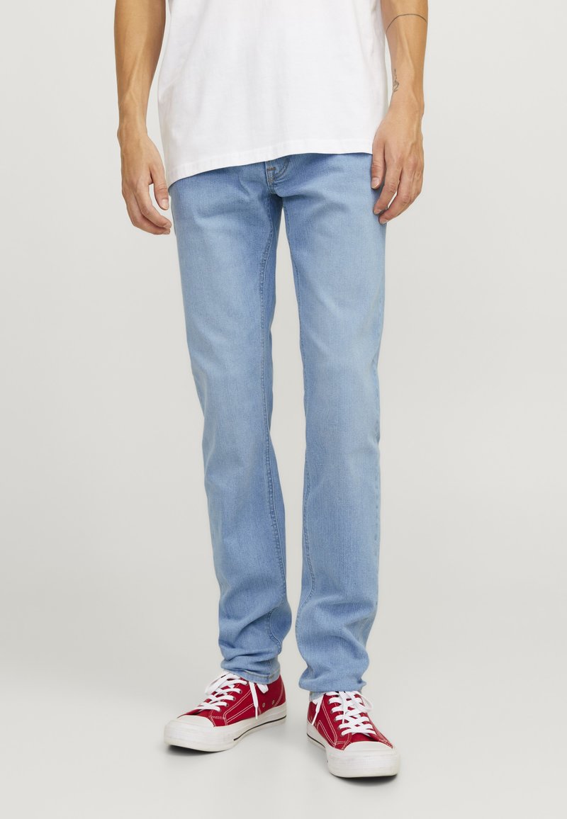 Jeans de mezclilla azul claro con corte recto, que presentan cinco bolsillos y una cremallera. Combinados con zapatillas rojas y una camiseta blanca.