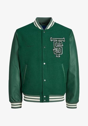 Man draagt een groene varsityjas met witte gestreepte boorden en kraag, donkerblauwe trui en lichtbeige broek, hand in de zak.