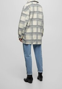 Wollen oversized jas in crème met grijze checks, voorzien van een opgestikte kraag, knoopmanchetten en een zachte textuur. In combinatie met blauwe jeans en zwarte laarzen.