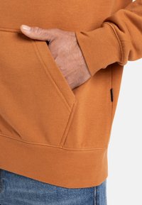 Orange bomullshuvtröja med en känguru ficka och ribbade ärmar, med en textur som är slät och mjuk, delvis visad på en modell.