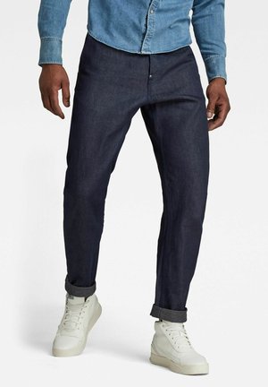 Homme portant un pantalon bleu foncé retroussé, une chemise en denim clair boutonnée et des baskets montantes blanches, debout devant un fond blanc.