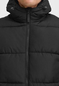 Giacca puffer nera con colletto alto, zip frontale e cappuccio regolabile con cordino. Il tessuto presenta una consistenza morbida e liscia con sezioni trapuntate.