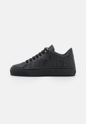 Zapatilla negra de corte bajo con un patrón sutil en relieve, cordones negros pasados por un ojete dorado y suela gruesa de goma sobre fondo blanco.