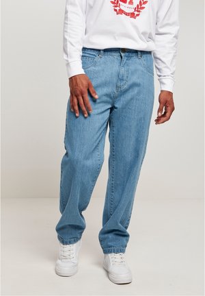 Una persona vestida con una camiseta blanca de manga larga y jeans azul claro está de pie con una mano en la cadera, mostrando un calzado informal.