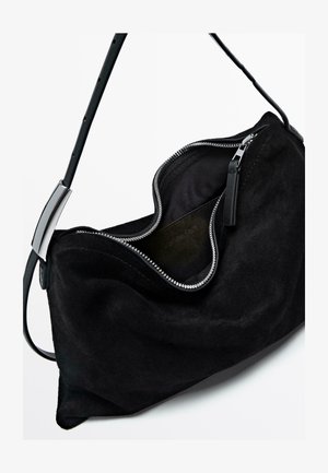 Sac à main en daim noir au design courbé, avec une fermeture éclair en argent et une fine bandoulière. L'intérieur est doublé de tissu foncé.