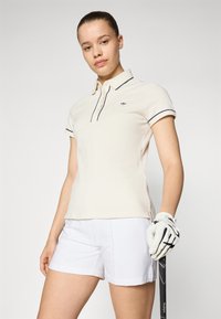 Krämfärgad pikétröja med marinblå detaljer, korta ärmar och liten logotyp. I kombination med vita shorts och håller en svart golfklubba.