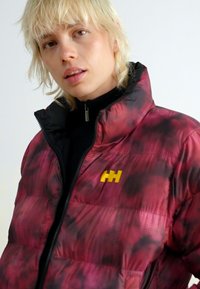 Chaqueta acolchada con un patrón rojo y negro, con un cuello negro y un logo amarillo en el pecho. El material parece brillante y acolchado.