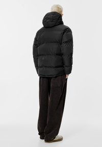 Črna puhasta jakna s kapuco, z vzorcem šivanke in debelo podlogo. Nošena z ohlapnimi, temno rjavimi hlačami in svetlimi čevlji.