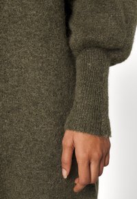 Oliivinvihreä villapaita, jossa on teksturoitu pinta ja leveät hihansuut. Kangas näyttää pehmeältä ja hieman pörröiseltä.