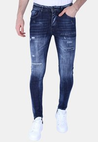Blauwe denim skinny jeans met een vervaagde wasbeurt, distressed details en meerdere scheuren; gedragen met witte sneakers en een aansluitend wit shirt.