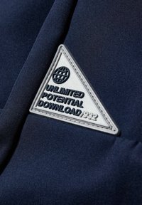 Granatowy materiał z trójkątną łatą z grafiką globu. Tekst wytłaczany czarnymi literami brzmi "NIEOGRANICZONY POTENCJAŁ POBIERZ 1932".