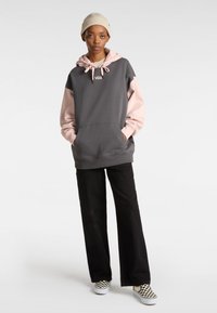 Sudadera bloqueada en color gris y rosa con un bolsillo delantero, capucha con cordones y un logo de Vans. Combinada con pantalones anchos negros y zapatos de cuadros.