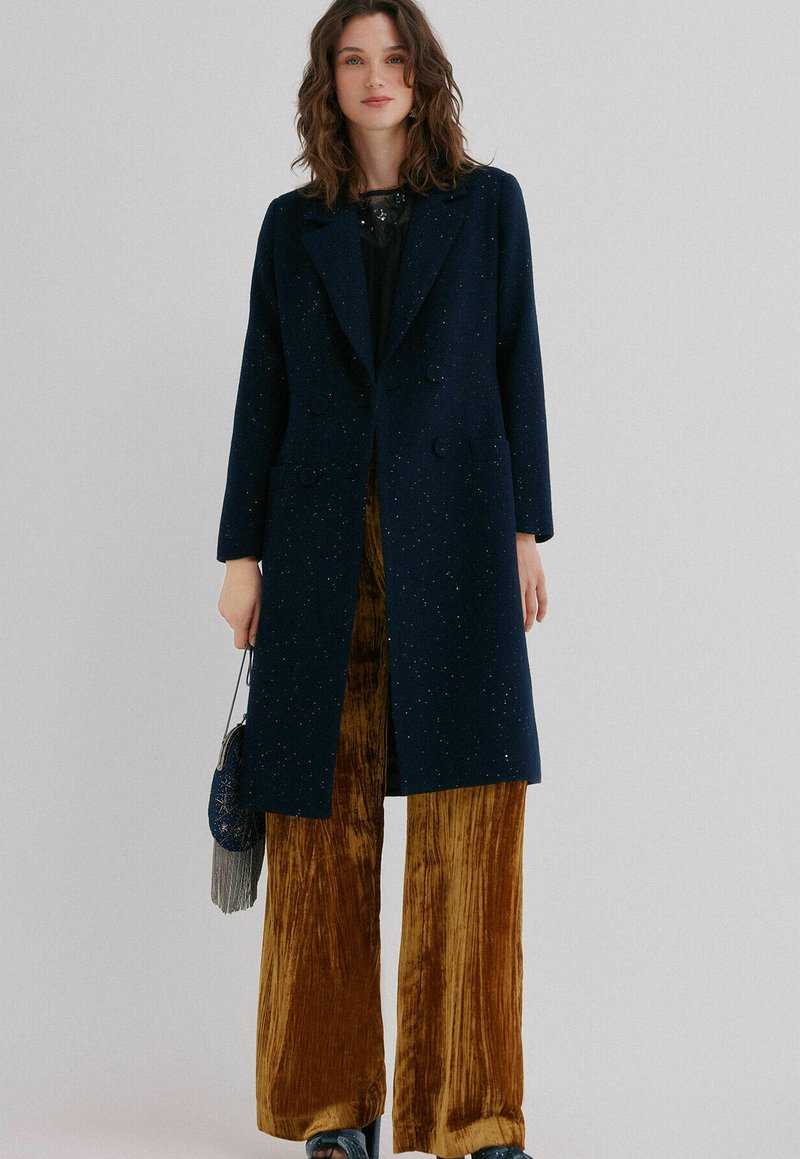 Femme portant un manteau croisé bleu marine foncé sur un haut noir et un pantalon velouté large moutarde, tenant un sac à main bleu marine avec des franges.