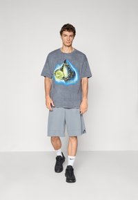 Ung mand står med en grå oversize T-shirt med troldmandsgrafik, grå knæ-lange shorts, hvide Nike sokker og sorte sneakers.