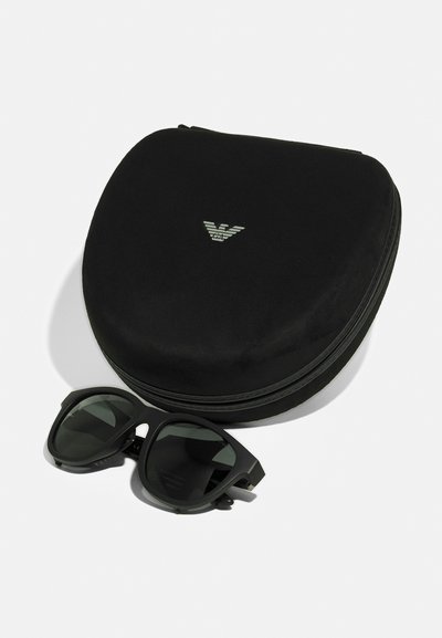 Occhiali da sole neri con lenti scure accanto a un astuccio rotondo e nero caratterizzato da un logo discreto. L'astuccio ha una texture liscia e una chiusura con zip.