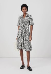 Abito a righe floreali bianco e nero con maniche corte, dettaglio in vita con cravatta e gonna fino al ginocchio. La modella tiene una borsa crema.
